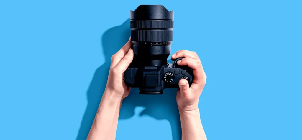 The biggest photography trends of 2025 2 Hands holding a black camera with a large lens against a blue background.