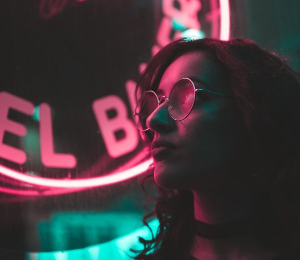 Woman with curly hair and glasses stands in front of a neon sign, casting a pink glow on her face.